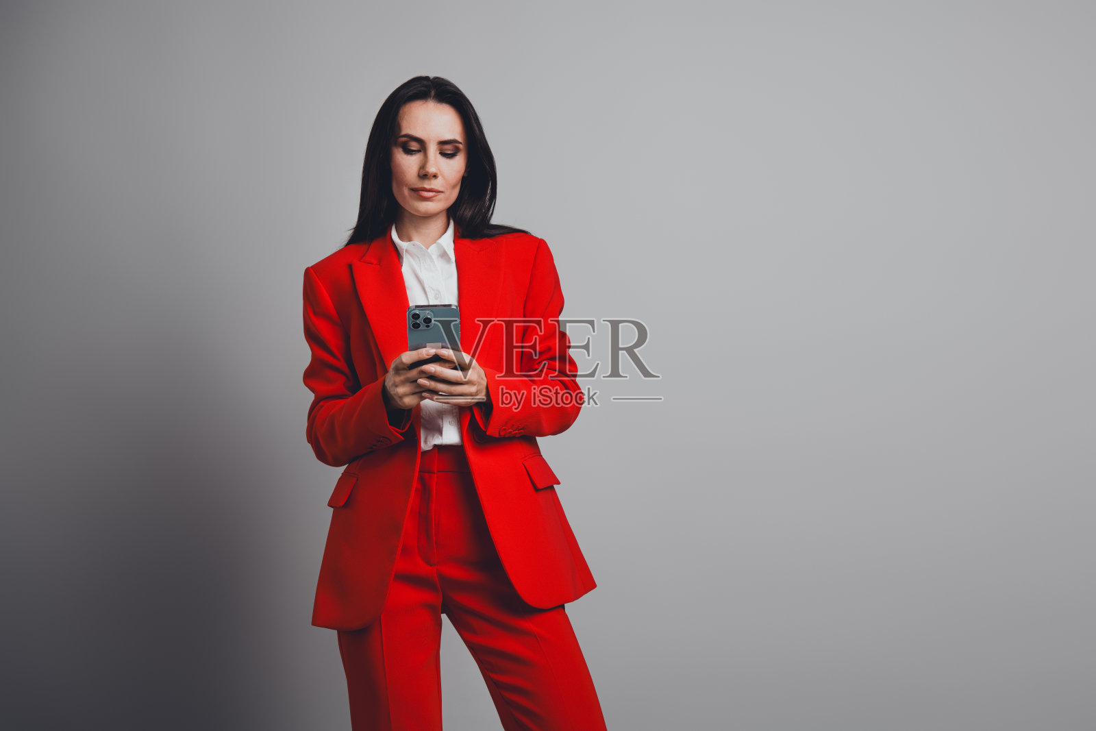自信的年轻女性专业人士穿着红色西装，优雅地摆姿势，手持智能手机，背景为灰色。照片摄影图片