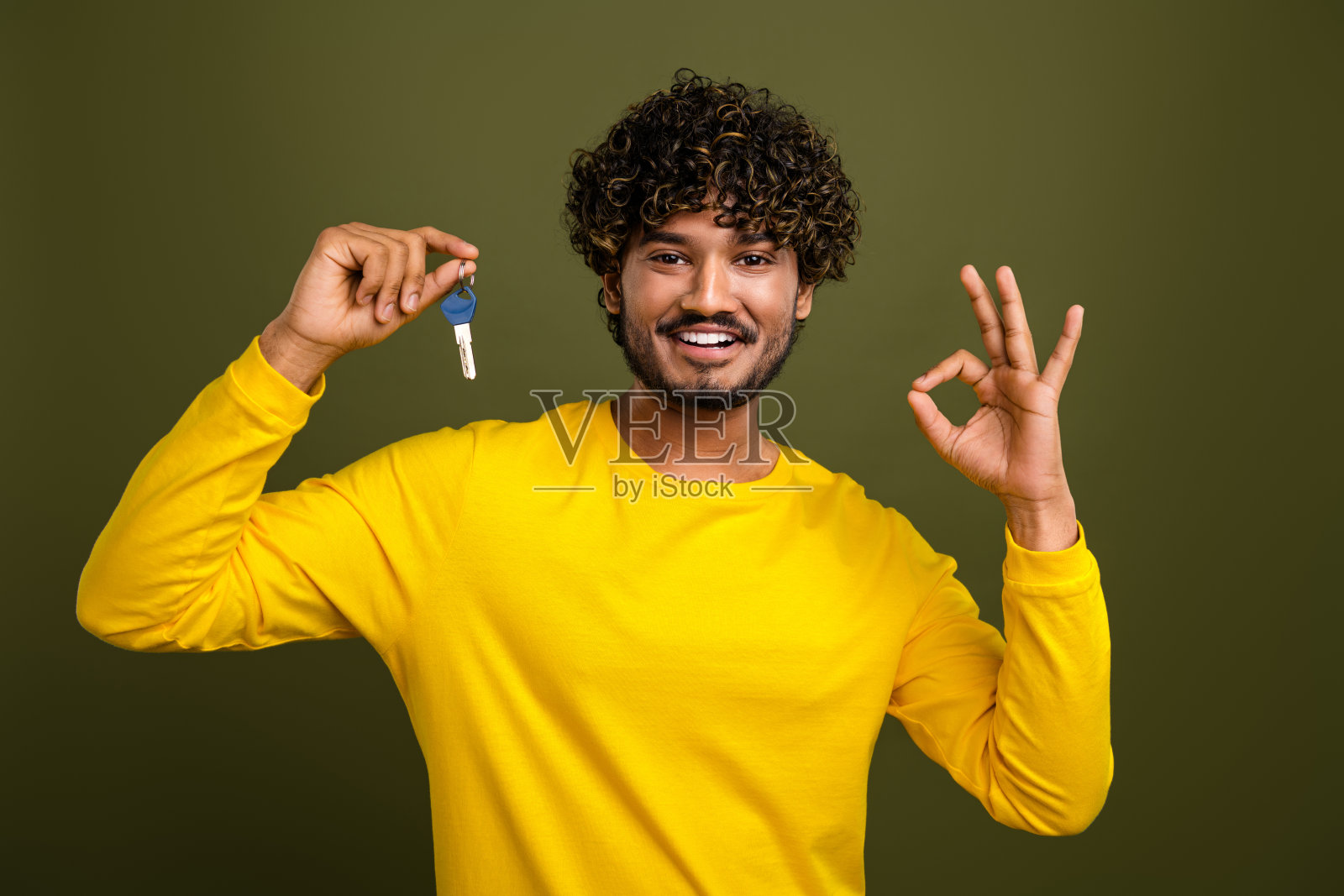 微笑的年轻男子手持房屋钥匙并在卡其色背景上做手势照片摄影图片