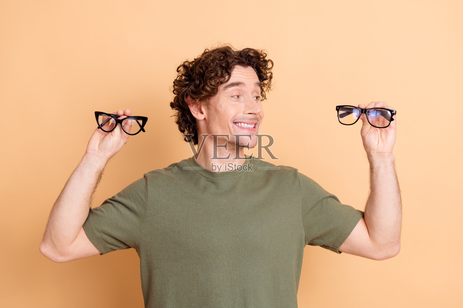 快乐的年轻男子穿着休闲服，双手各持一副时尚眼镜，背景为米色。照片摄影图片