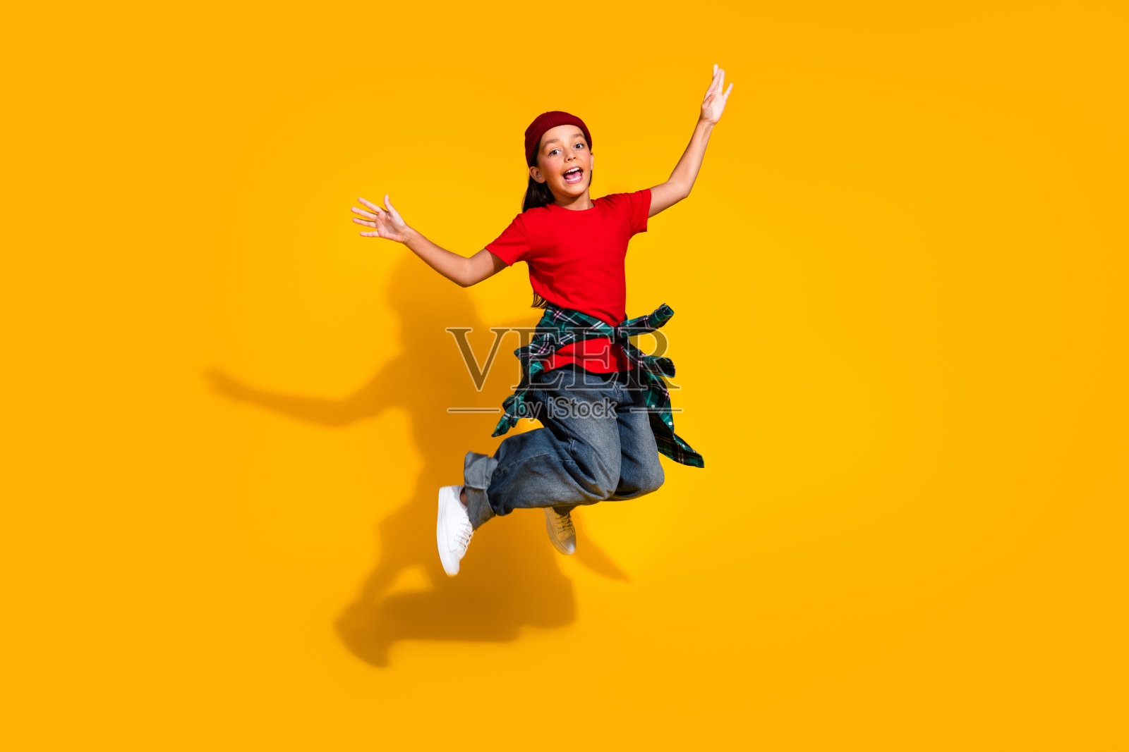 充满活力的年轻女孩在鲜艳的黄色背景上跳跃，穿着时尚休闲的服装，戴着毛线帽和格子衬衫。照片摄影图片