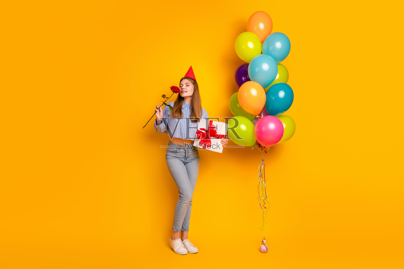 年轻女性在明亮的黄色背景下庆祝，手持气球、礼物和节日帽子照片摄影图片