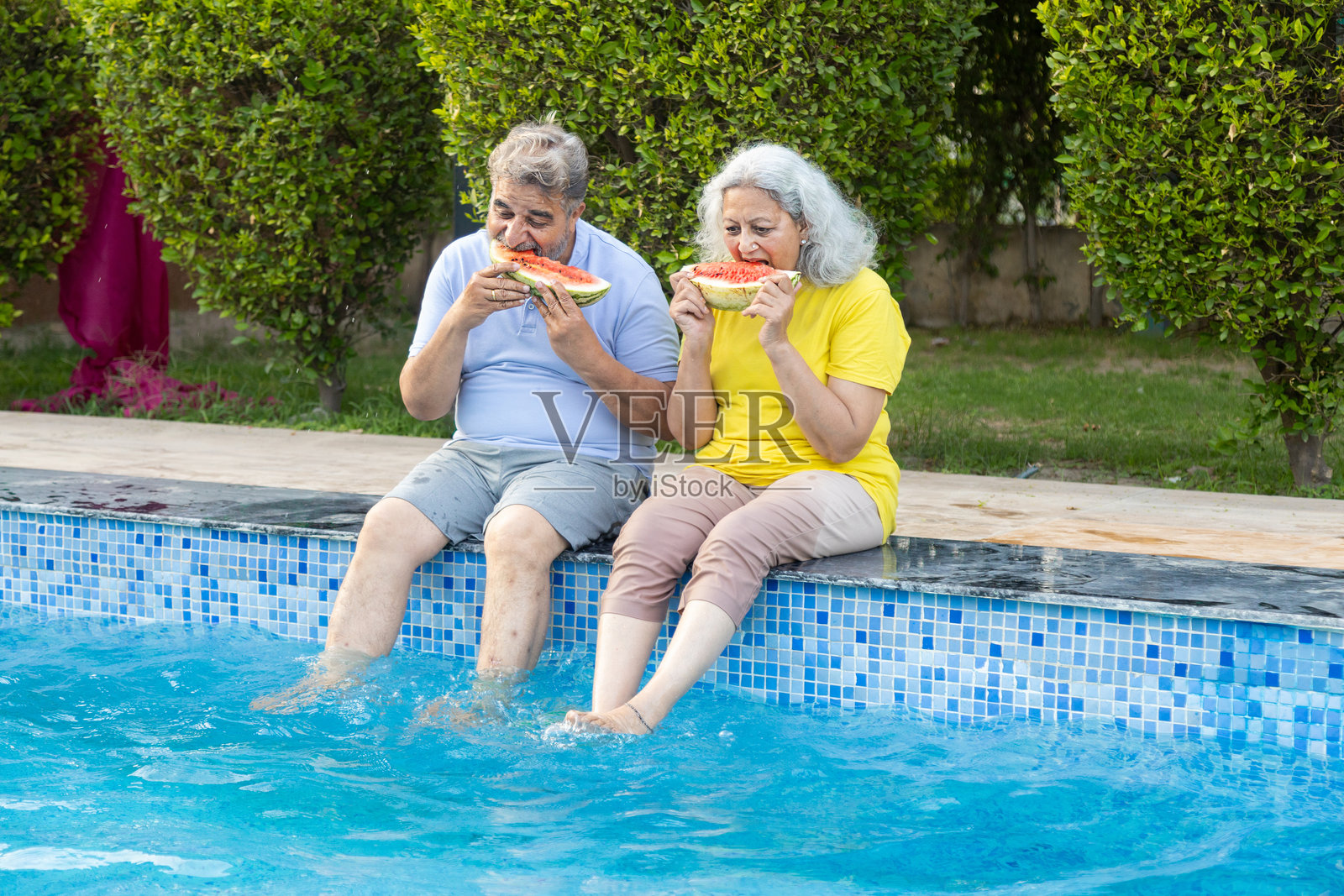 夏季假期 - 一对退休的印度老年夫妇在游泳池享用西瓜，成熟的退休人士在度假酒店放松享受乐趣。退休生活。亲密关系与情感纽带。照片摄影图片