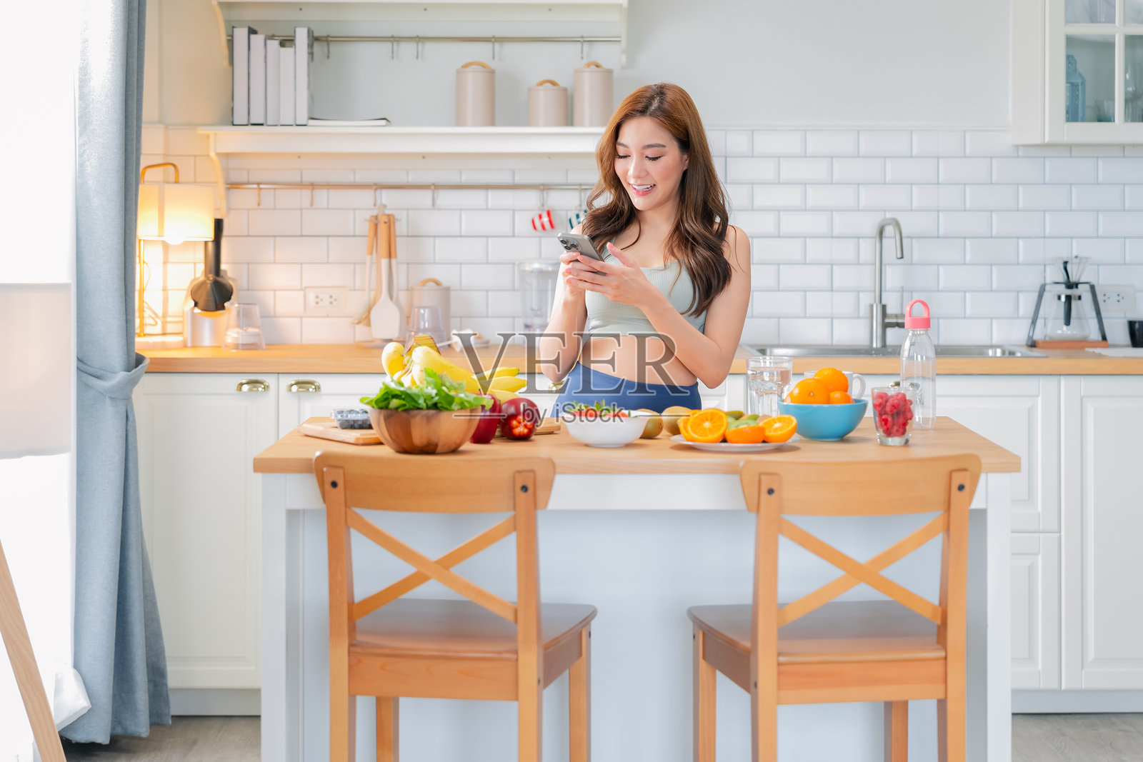 身穿时尚运动服的亚洲美女在现代家居厨房中准备健康的水果饮品，微笑着用智能手机拍照，准备分享在社交媒体上，展现健康生活方式。照片摄影图片