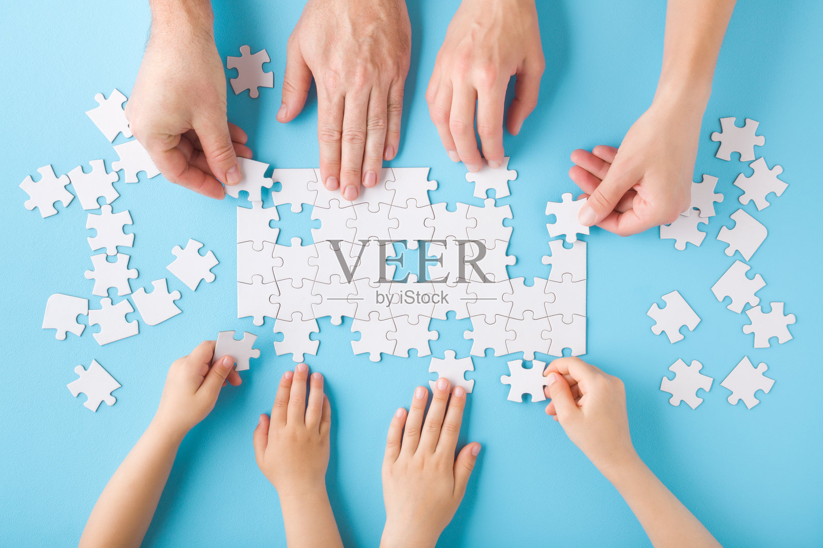 母亲、父亲和孩子们的手在浅蓝色桌面上玩耍并拼装白色拼图块。柔和的色彩。家庭活动。共度时光。特写。俯视图。照片摄影图片