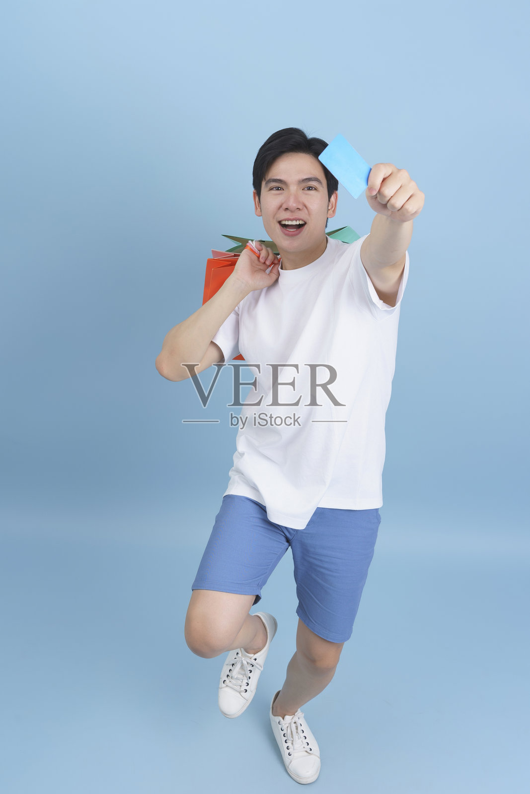 兴奋的年轻男子手持信用卡，身旁有购物袋，背景为蓝色照片摄影图片