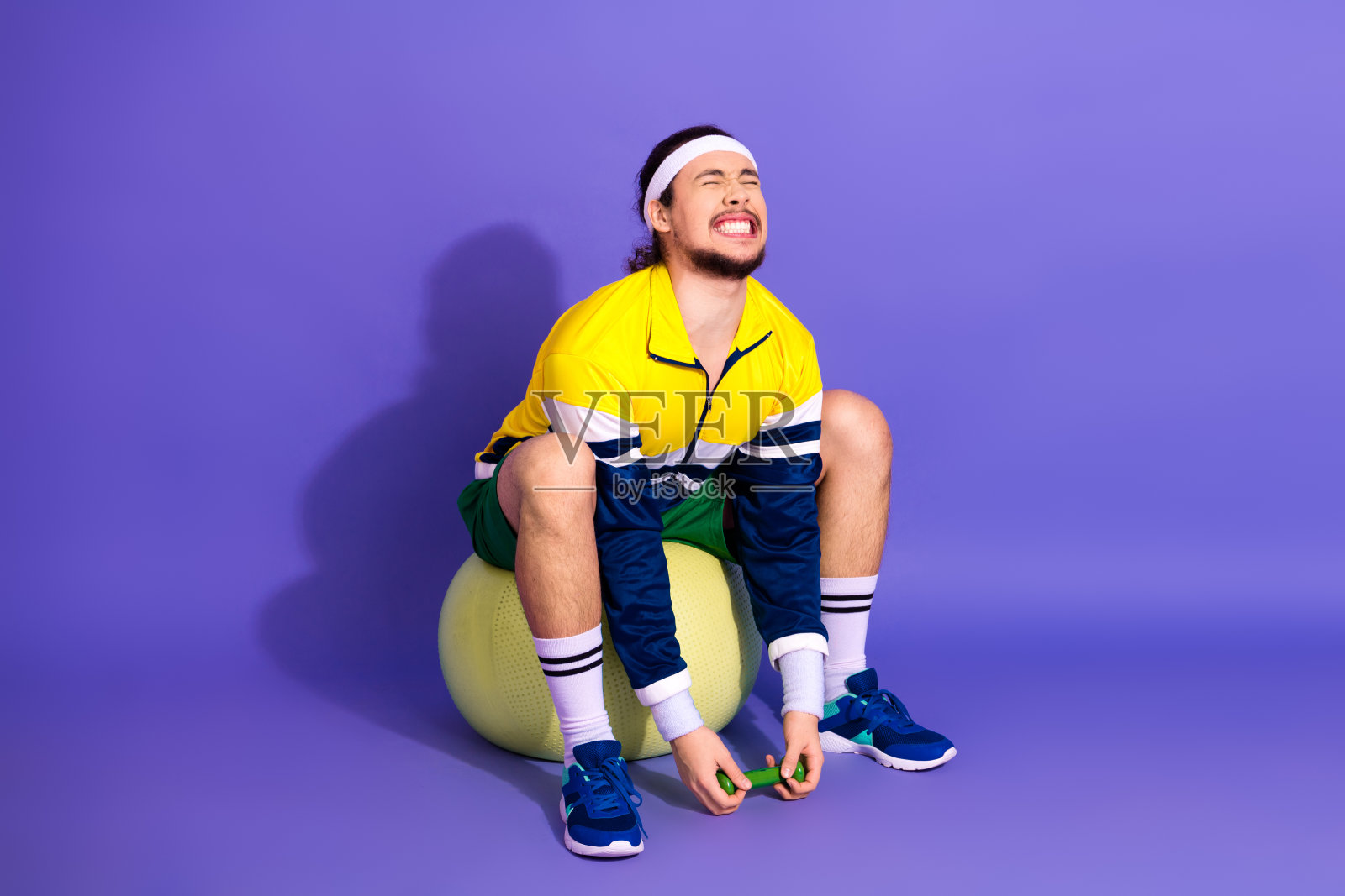 活力男士在色彩缤纷的健身房环境中，穿着复古运动服，使用小哑铃在健身球上锻炼。照片摄影图片