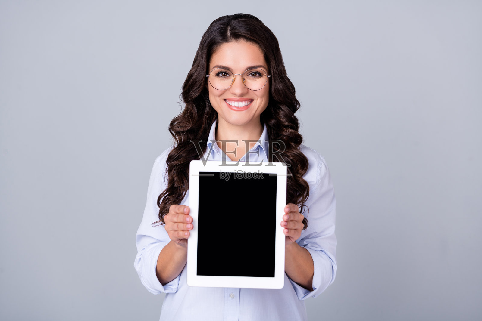 一位积极快乐的女士戴着眼镜，手持平板电脑，空白屏幕，穿着蓝色衬衫，背景为灰色。照片摄影图片