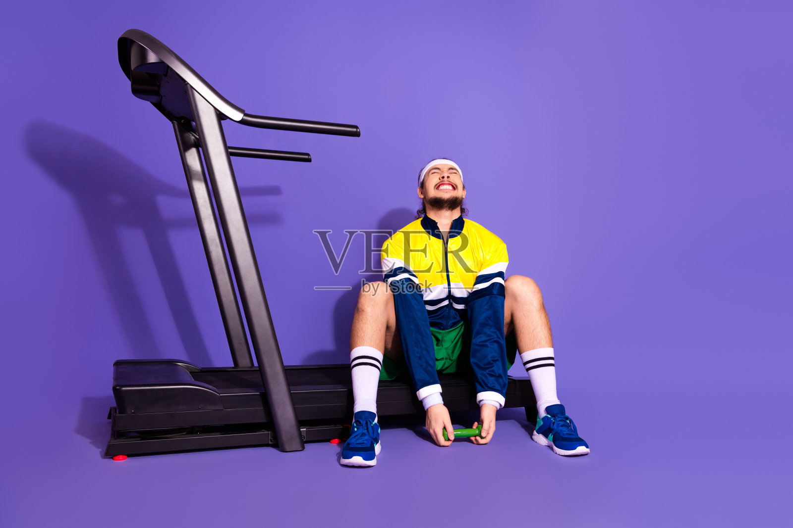 年轻男性运动员穿着复古运动服，愉快地坐在跑步机旁，背景为鲜艳的紫色。照片摄影图片