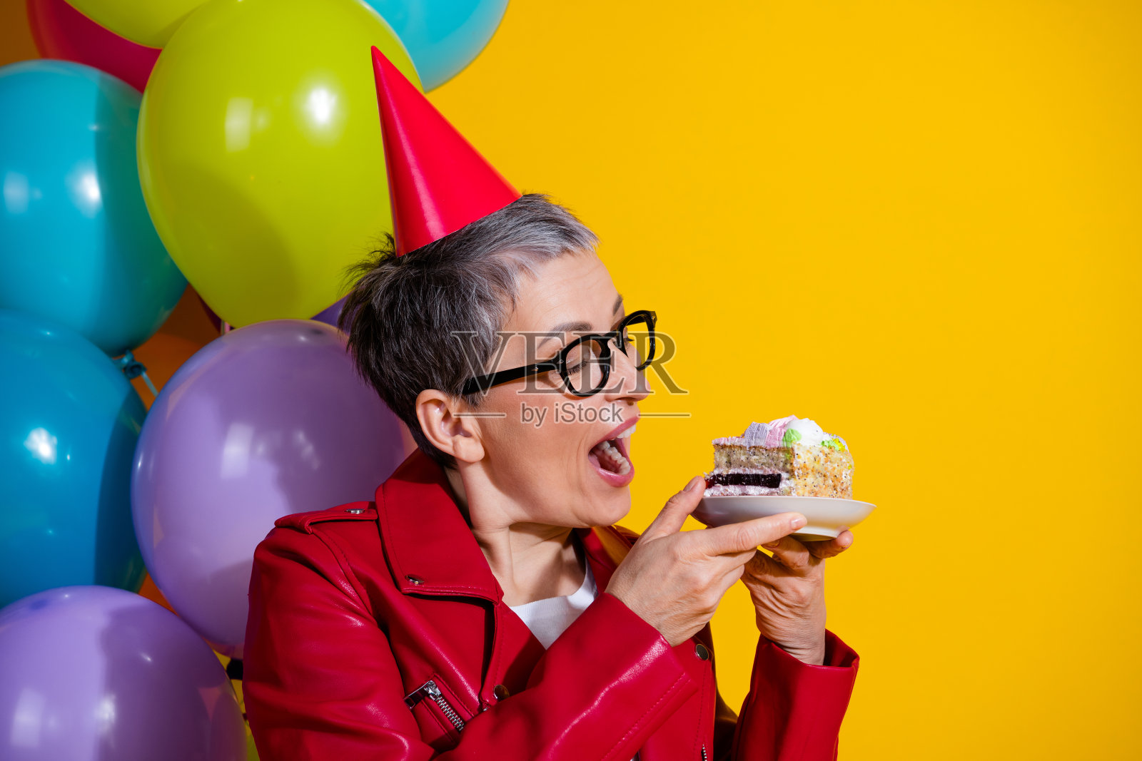 快乐的女性穿着节日派对服装，享受五彩缤纷的蛋糕，周围环绕着气球，背景是鲜艳的黄色。照片摄影图片