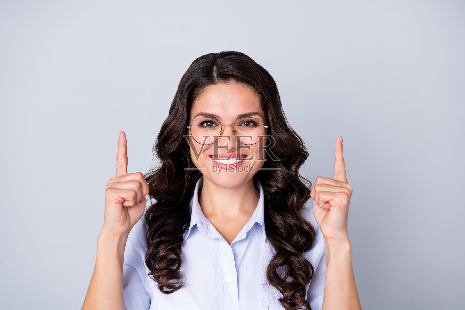 年轻的棕发波浪卷女教师，戴着眼镜，手指竖起，穿着蓝色衣服，背景为灰色。照片摄影图片