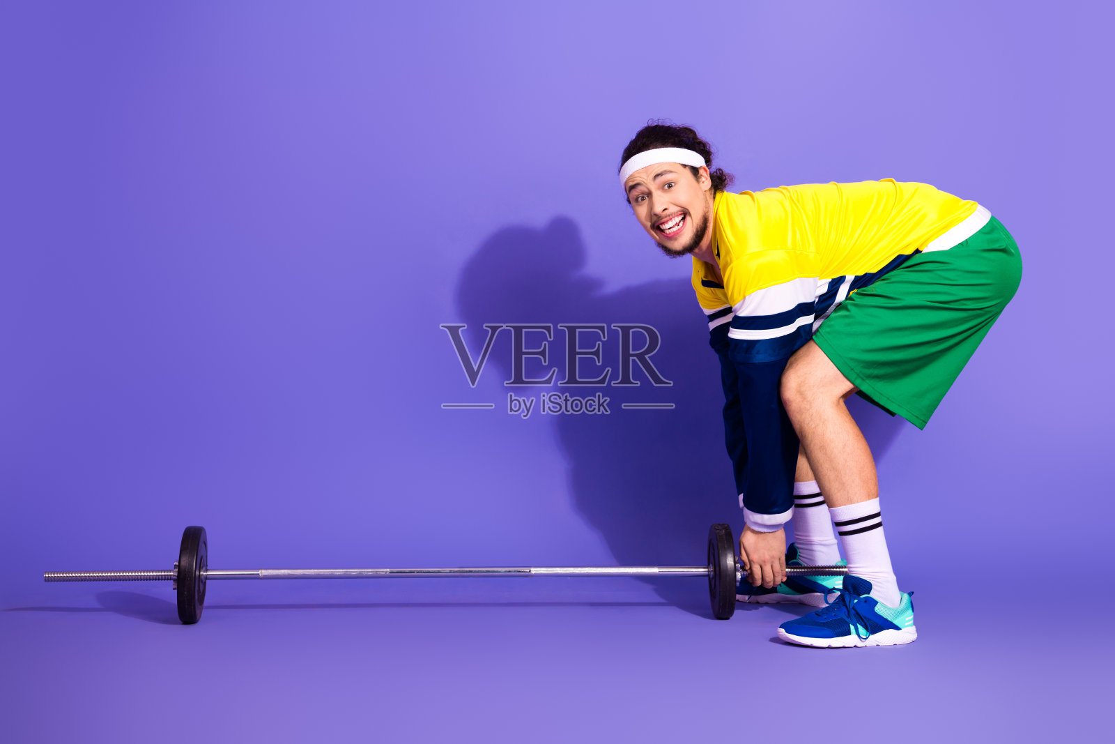 年轻男子穿着复古运动服，在紫色背景下愉快地举着杠铃，展现出复古风格。照片摄影图片