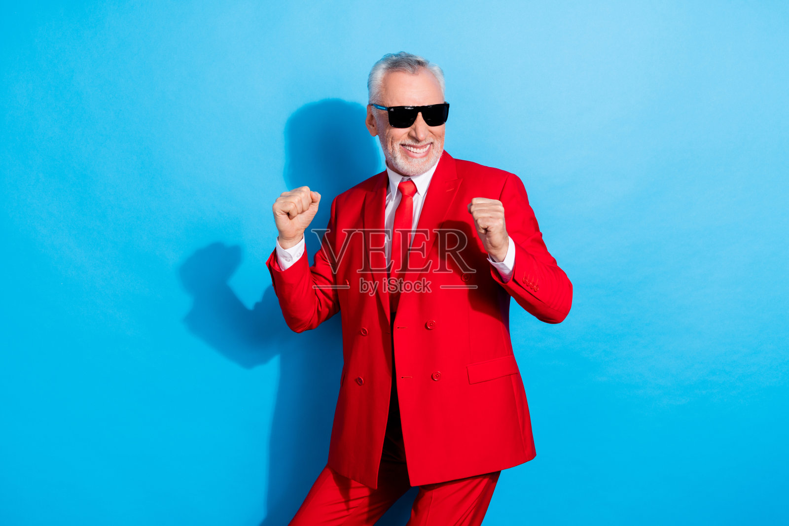 穿着红色礼服、戴着太阳镜的快乐退休男士在蓝色背景下享受迪斯科舞蹈。照片摄影图片