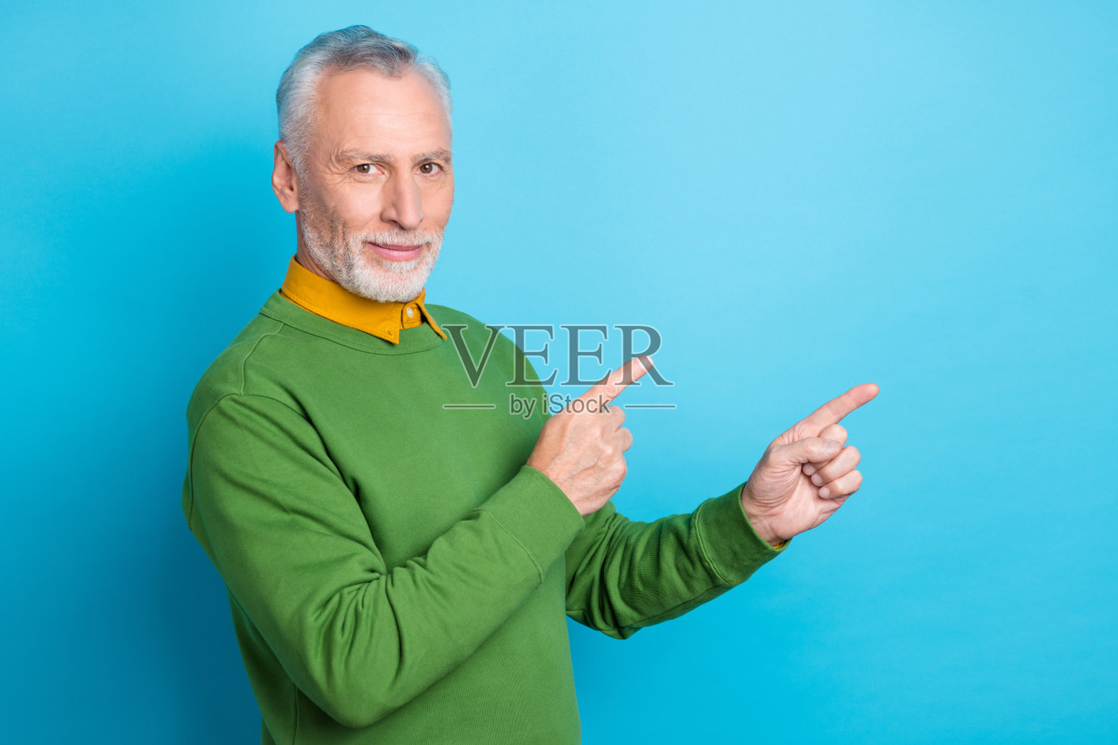 穿着绿色毛衣的积极退休男性，双手指向空旷的地方，背景为蓝色。照片摄影图片