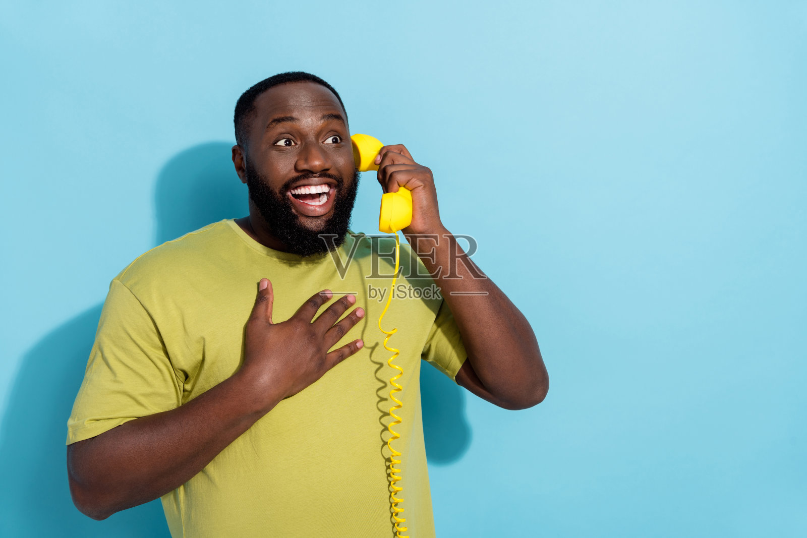 穿着绿色T恤的非洲裔男士，手臂交叉，胸前有些惊讶，正在使用复古电话，背景为空旷的蓝色。照片摄影图片