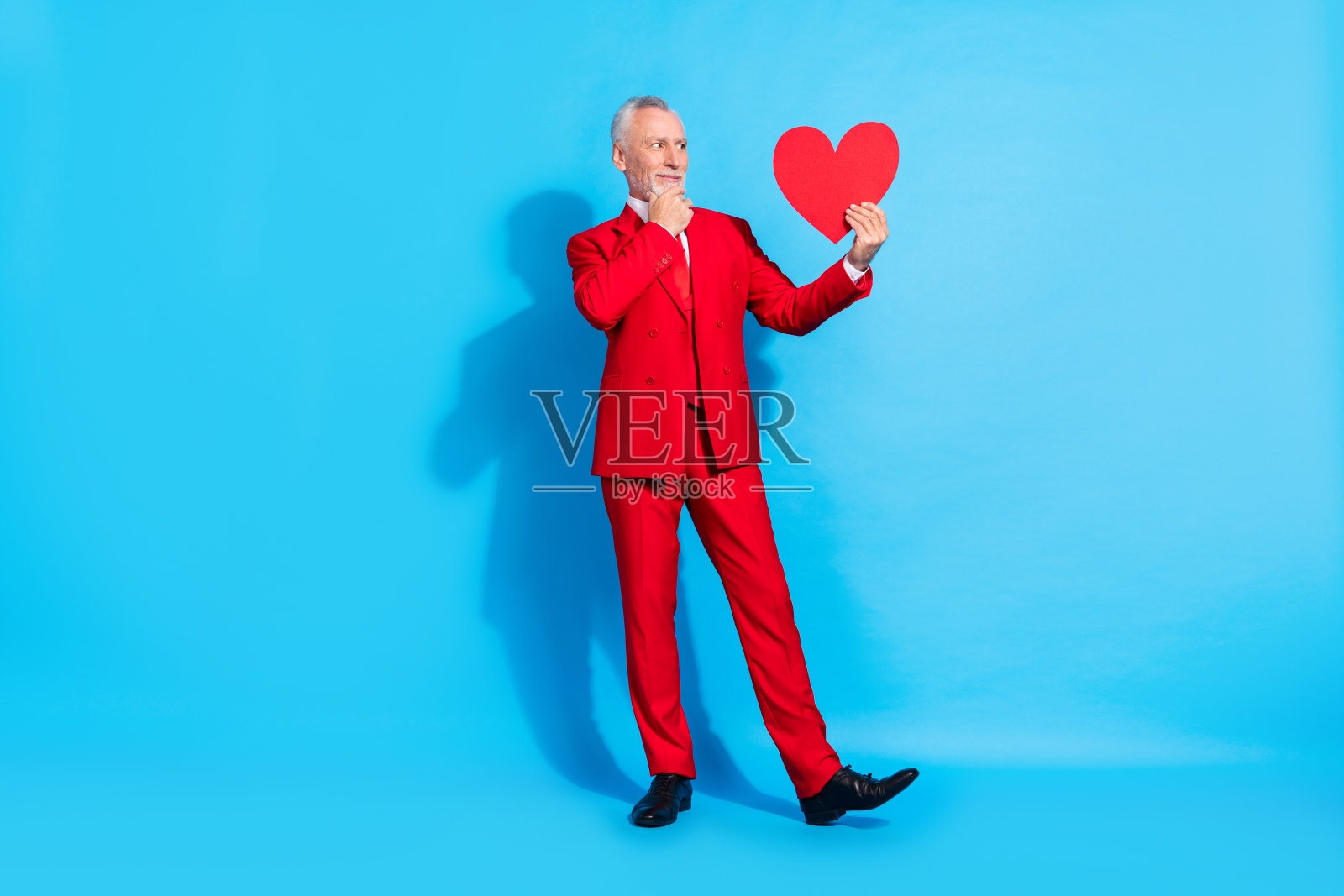 穿着红色西装的聪明男友，手持心形卡片，手指托着下巴，思考中，背景为蓝色。照片摄影图片