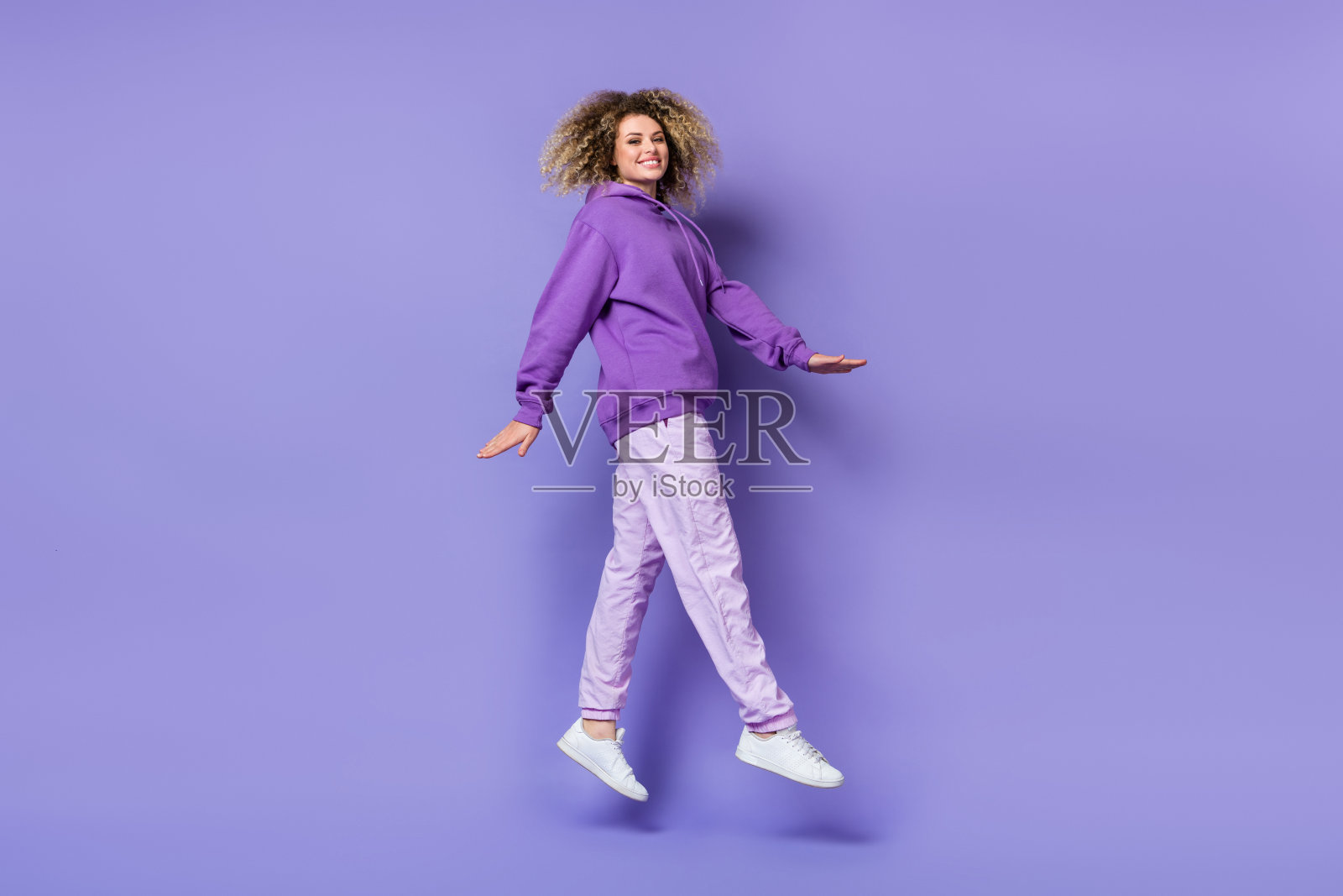 穿着卫衣的甜美兴奋女性在紫色背景下高高跳起。照片摄影图片