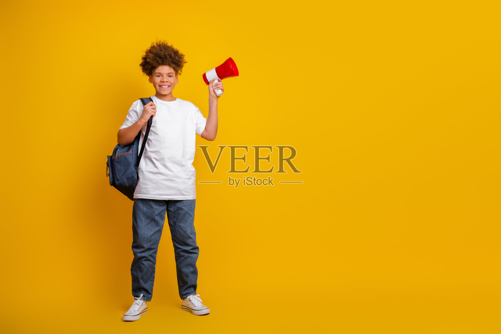 微笑的卷发少年手持扩音器，穿着休闲风格的衣服，站在鲜艳的黄色背景前照片摄影图片