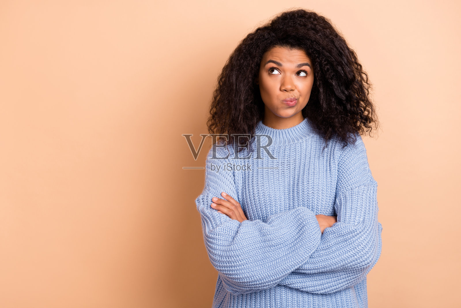 年轻女性双手交叉，思考着，目光疑惑，面对空旷的空间，孤立在米色背景前。照片摄影图片