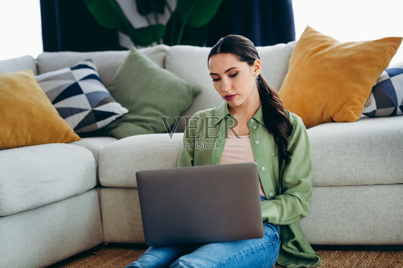 年轻女性坐在家中舒适的客厅里，使用笔记本电脑，周围环绕着五颜六色的抱枕。照片摄影图片