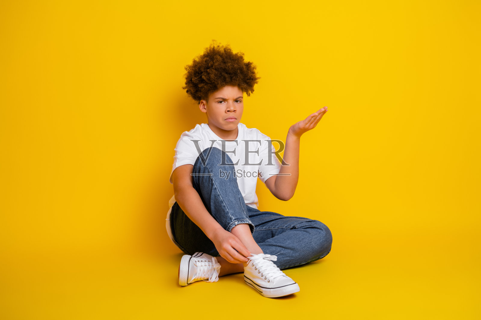 年轻男孩坐在黄色背景前，做出富有表现力的手势，穿着休闲服和运动鞋，展现生活方式概念照片摄影图片