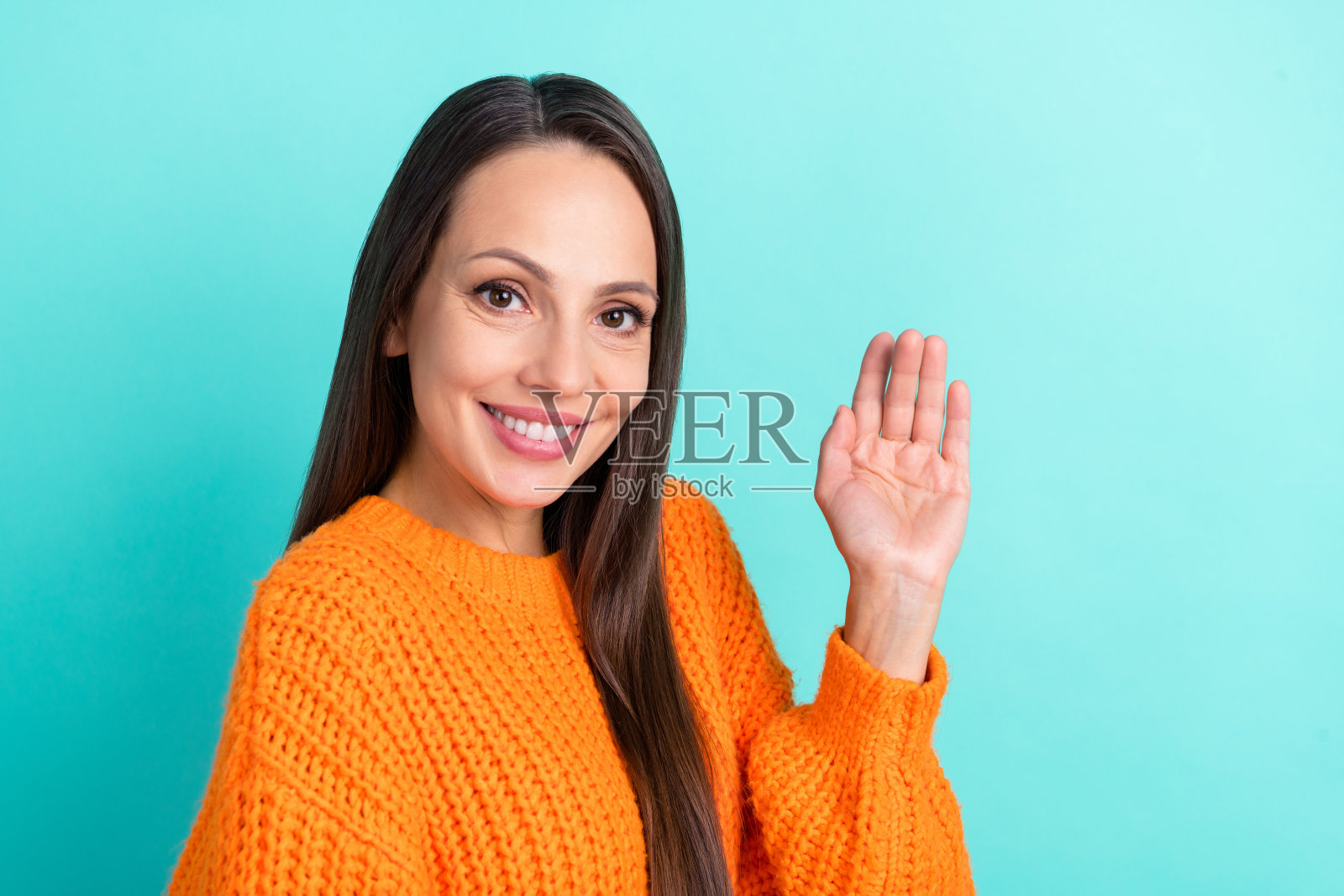 可爱的年轻棕发女孩穿着橙色毛衣，站在青绿色背景前微笑挥手。照片摄影图片