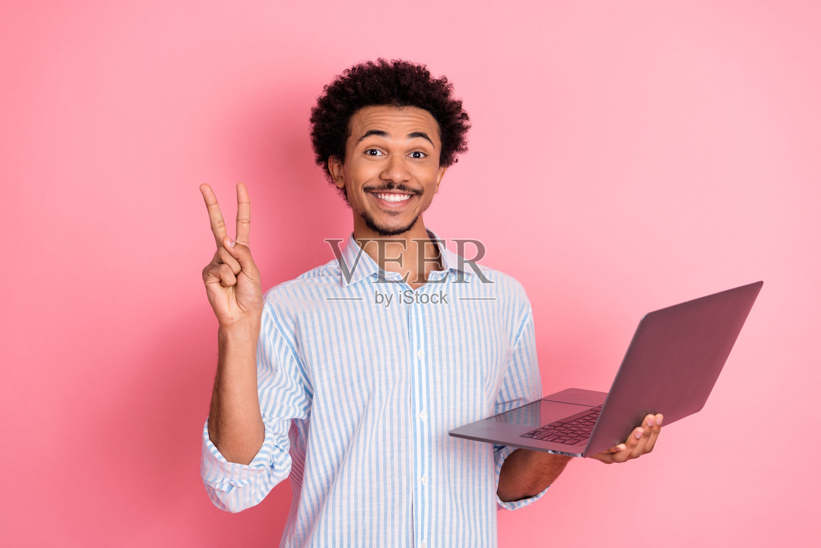 微笑的年轻男子手持笔记本电脑，做出和平手势，背景为粉色，穿着休闲服，表达兴奋之情照片摄影图片