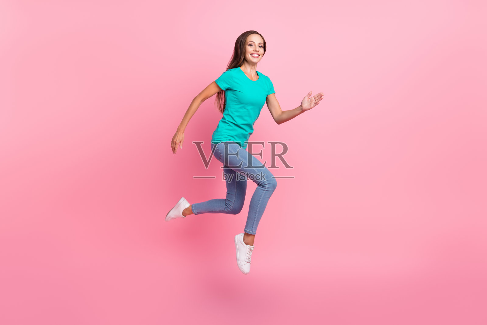穿着青色T恤和牛仔裤的活力四射的女士在粉色背景上跳跃向前奔跑的照片照片摄影图片