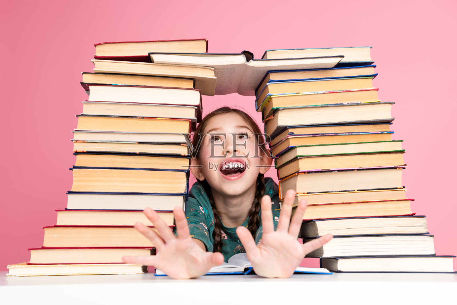 可爱的女孩在粉色背景下兴奋地伸手，周围是堆积如山的书籍。照片摄影图片