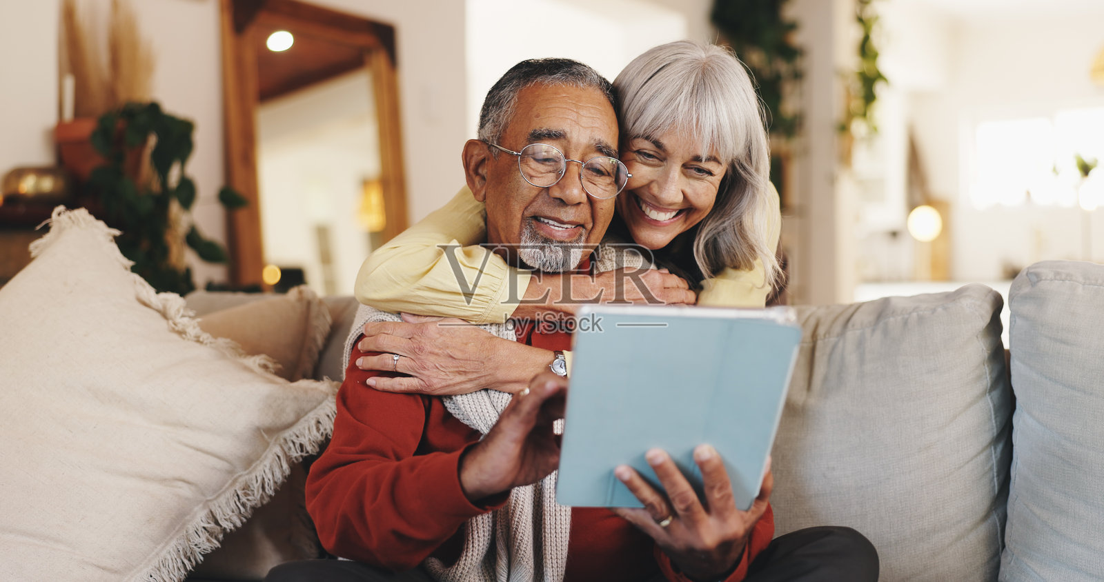 家中，平板电脑和一对老年夫妇相拥，放松地交谈，使用应用程序，充满爱意与互联网。人们、关系，以及老年男性与老年女性，科技与娱乐，社交媒体与拥抱。照片摄影图片