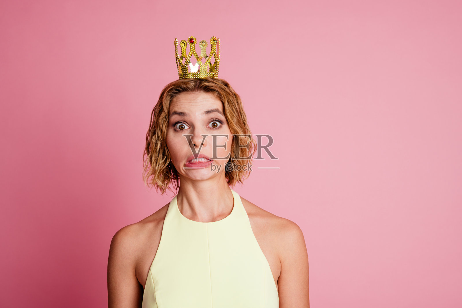 年轻女性戴着皇冠，在粉色背景前做出搞笑表情，展现出魅力和俏皮风格照片摄影图片