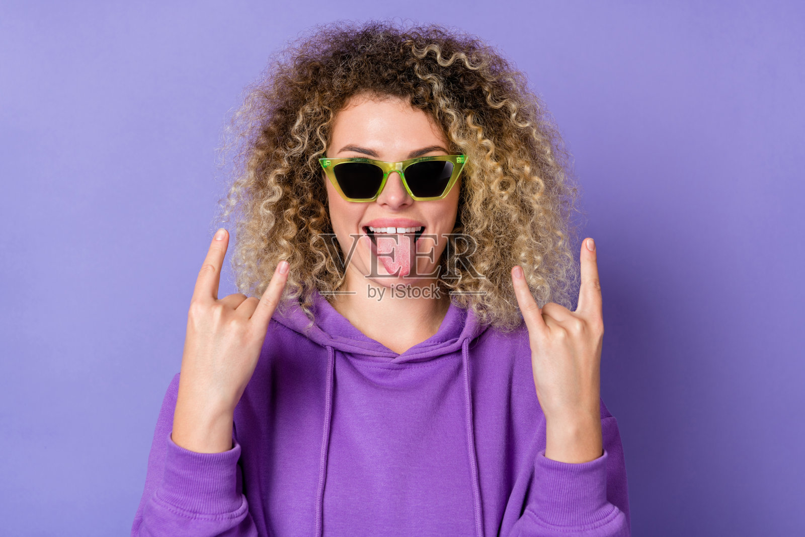 穿着卫衣、戴着深色眼镜的酷炫角女人，做出摇滚手势，舌头伸出，背景为紫色。照片摄影图片