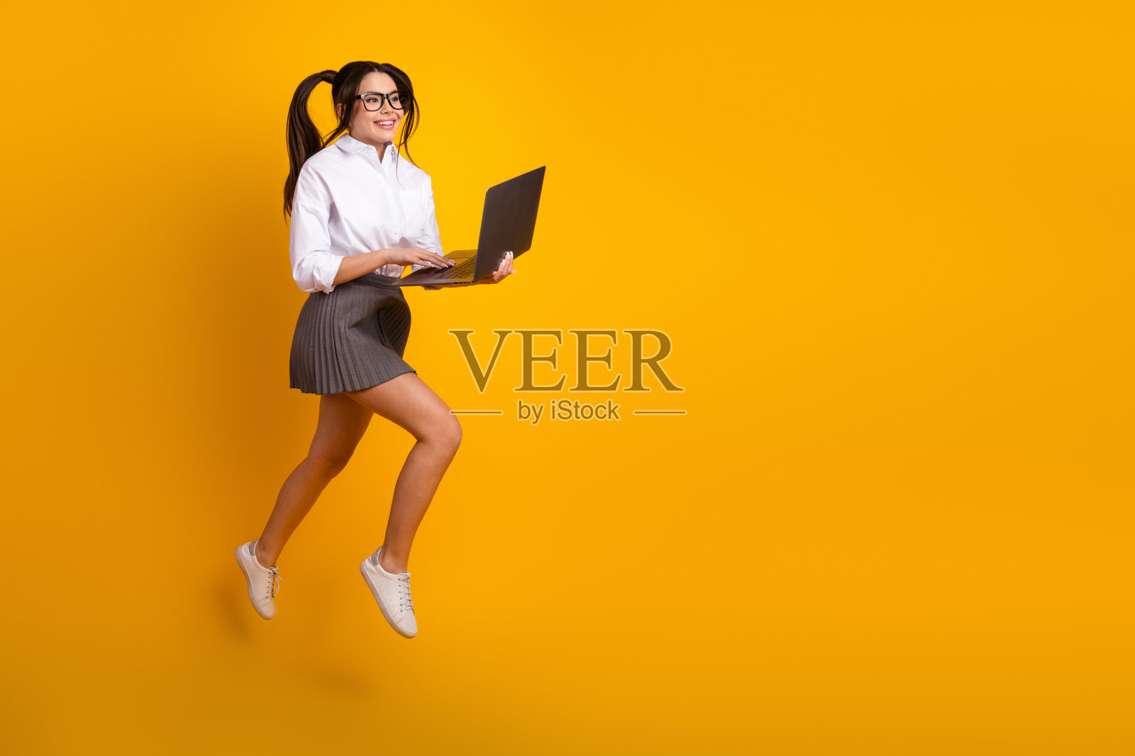 年轻女性在充满活力的黄色背景上跳跃，穿着休闲的校园服装，散发着快乐和活力。照片摄影图片