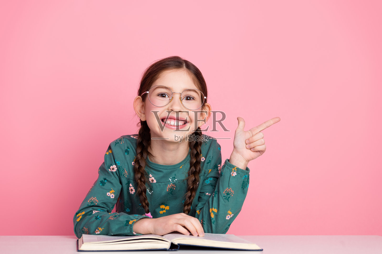 微笑的辫子女孩在粉色背景下兴奋地摆姿势，手中拿着一本打开的书。照片摄影图片