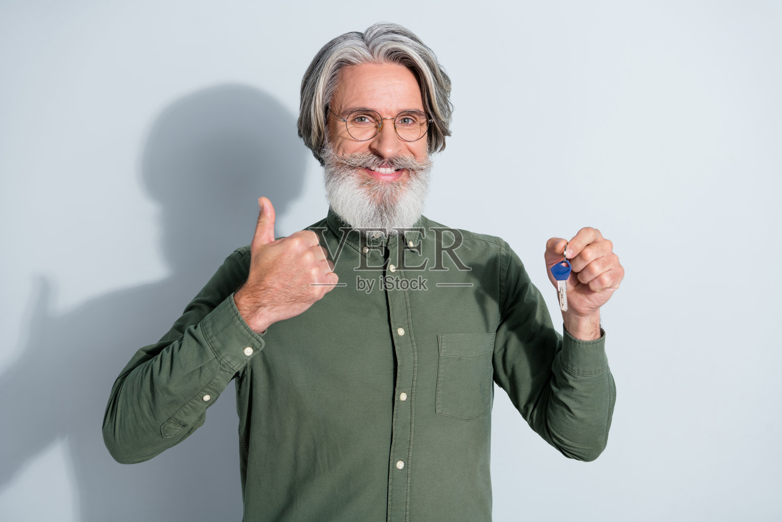 自信的退休男士穿着卡其色衬衫，戴着眼镜，手握钥匙，竖起大拇指，背景为灰色。照片摄影图片