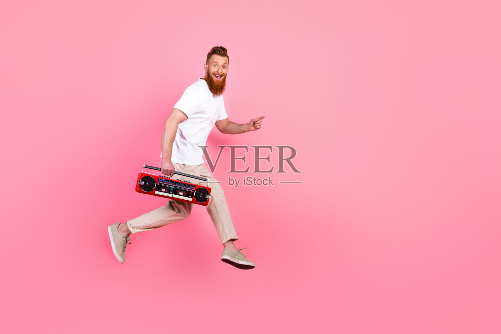 年轻时尚男子穿着休闲服，手持红色复古音响，正处于动态中，背景为明亮的粉色。照片摄影图片