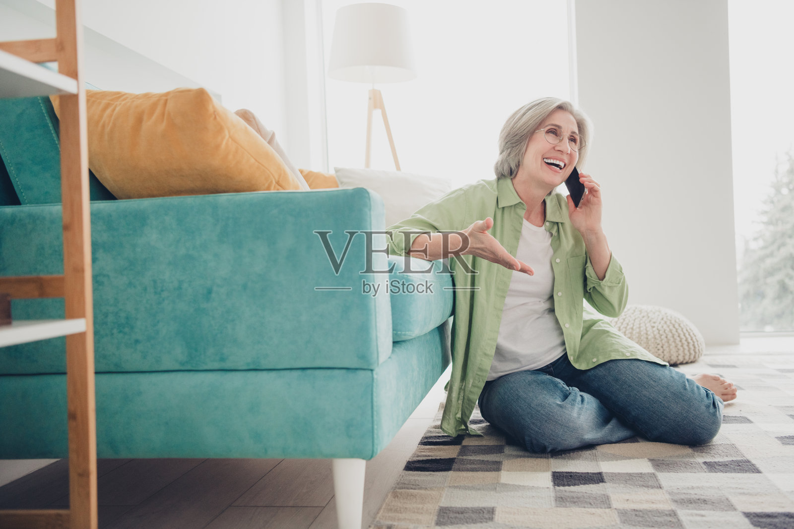 一位老年女性坐在地板上，靠在浅蓝色沙发上，在温馨明亮的室内打电话。照片摄影图片
