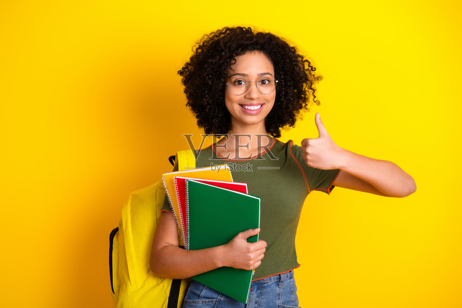 自信的年轻学生在明亮的黄色背景下，手持五彩缤纷的笔记本，竖起大拇指。照片摄影图片