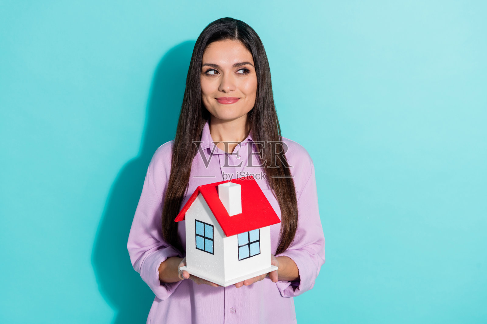 梦幻般的房地产女代理人，手持房屋模型，穿着紫色衬衫，站在空旷的空间中，背景为青色。照片摄影图片