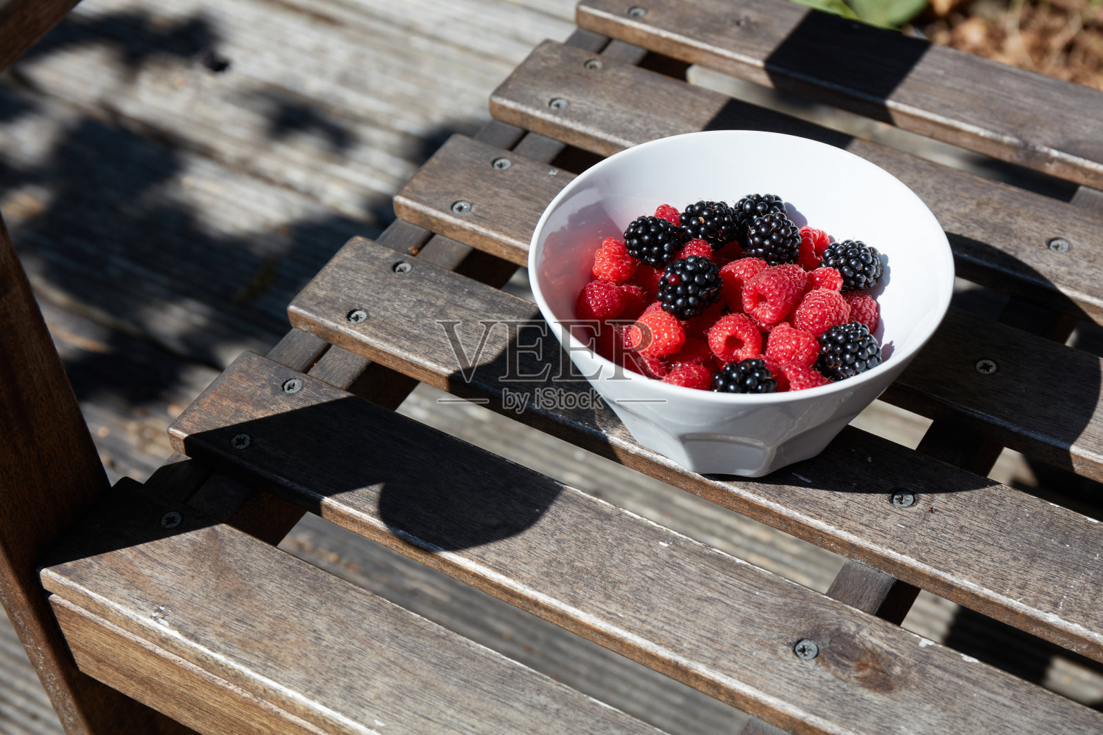 鲜艳的覆盆子和黑莓装满了一个白色碗，碗放在阳光明媚的花园中的木桌上。水果在自然光下展现出明亮的色彩。照片摄影图片