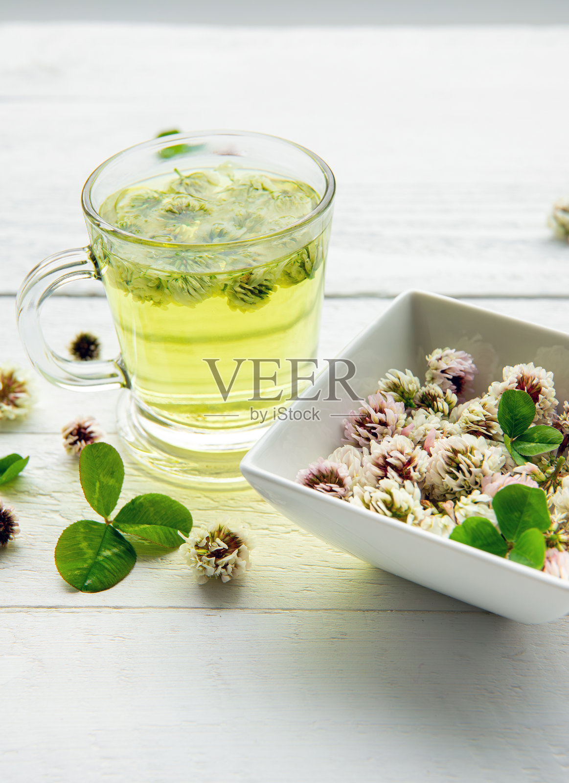 白三叶草（Trifolium repens）草本茶在透明玻璃杯中。热气腾腾的饮品中有花朵，旁边是一个碗，背景为简约的白色木板。照片摄影图片