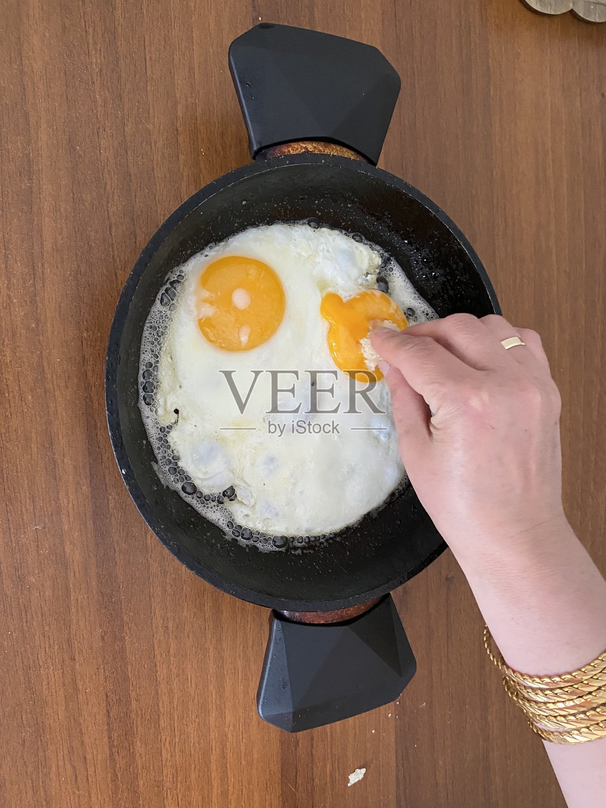 一位白人女性的手。她用面包戳破两个煎蛋中的一个蛋黄。照片摄影图片