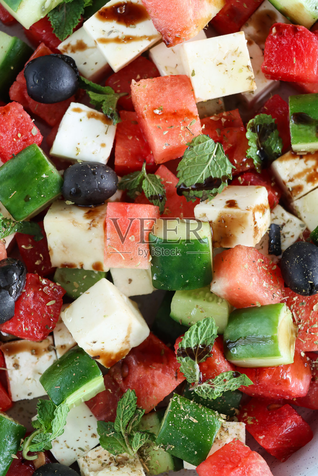 希腊西瓜沙拉的全景图，碗中盛有切片黄瓜、红洋葱、费塔奶酪块、薄荷叶和黑橄榄，淋上香醋，蓝色背景，前景聚焦，健康饮食概念照片摄影图片
