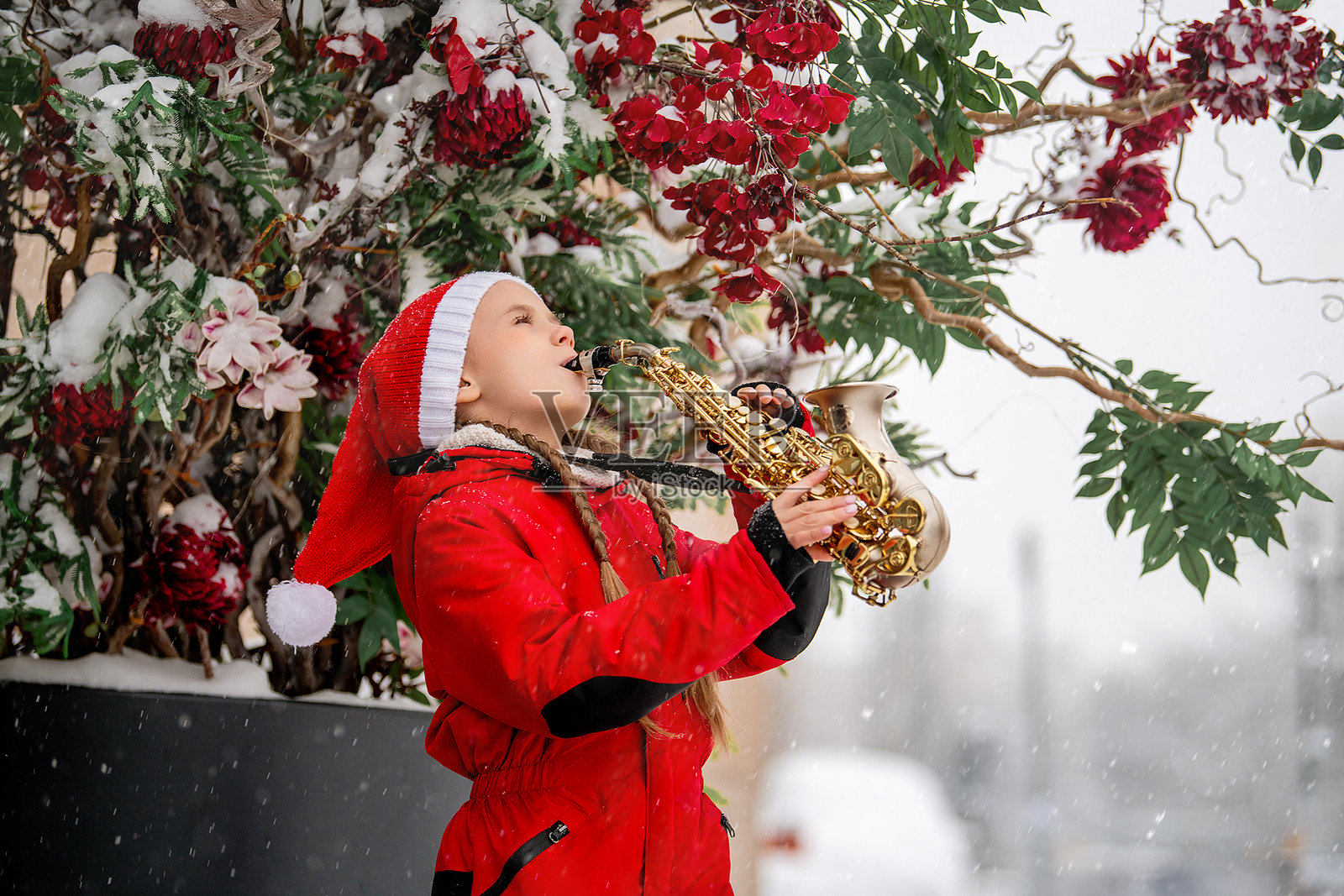 穿着红色圣诞老人帽的小女孩在城市街道上雪中演奏萨克斯ophone。照片摄影图片