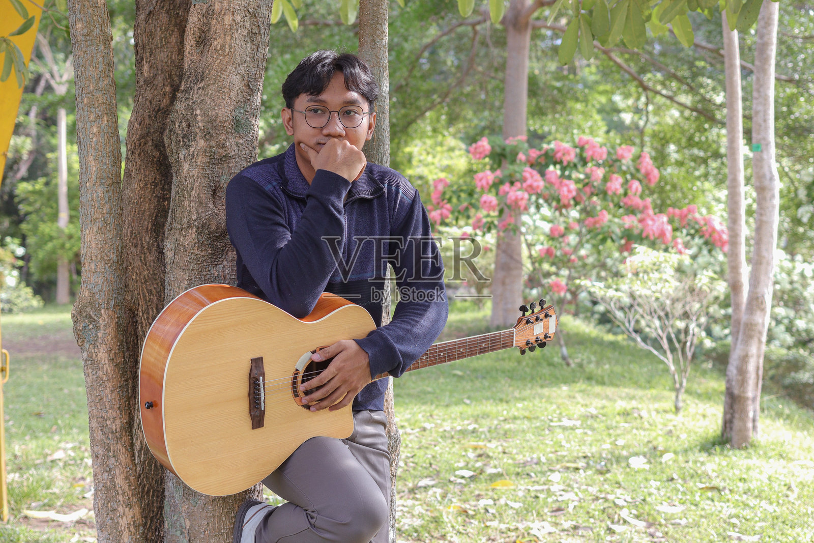 亚洲男子在城市公园的温暖夏日中弹吉他，四周环绕着绿色植物。宁静的氛围、明亮的阳光和悦耳的旋律营造出一个平和而充满活力的时刻。照片摄影图片