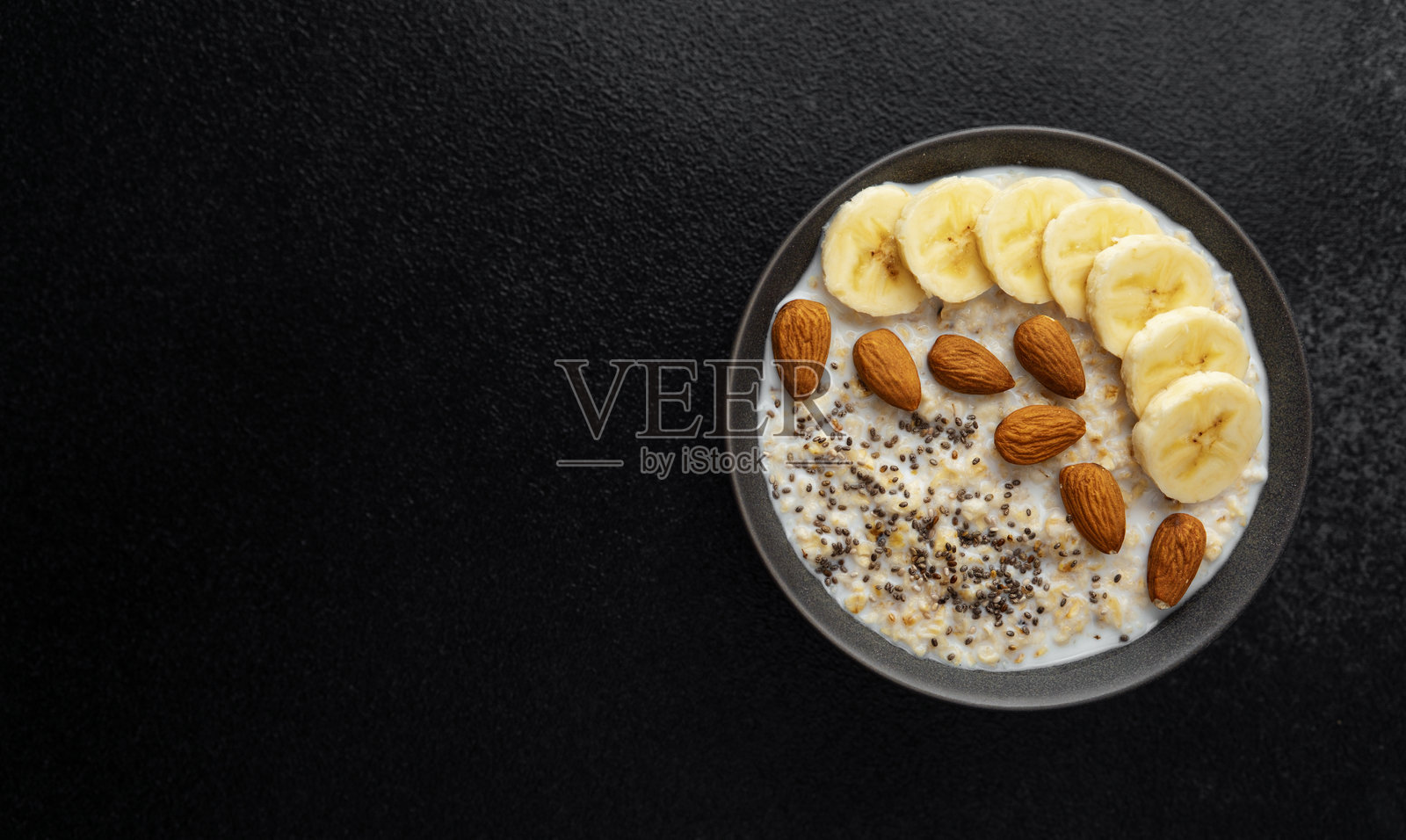 黑色背景下，深碗中盛着燕麦粥，配有香蕉片、杏仁和奇亚籽。照片摄影图片