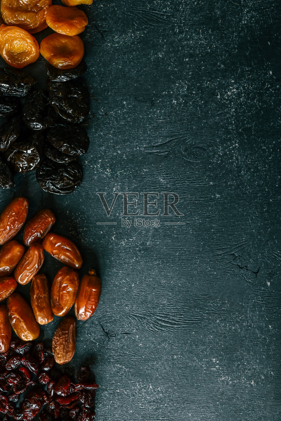 四种干果——蔓越莓、枣、梅干和杏干——在深色纹理背景上排列成行。色彩丰富且健康的天然零食组合。照片摄影图片