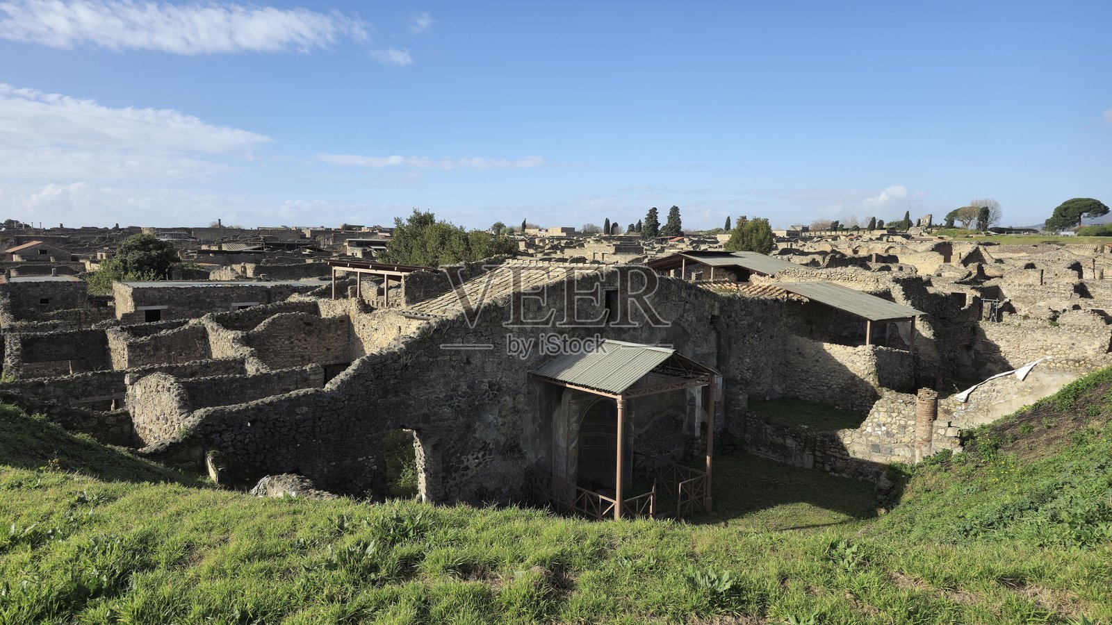 庞贝遗址的宽阔视野，屋顶结构保护着关键区域，背景是晴朗的天空和草地前景。照片摄影图片