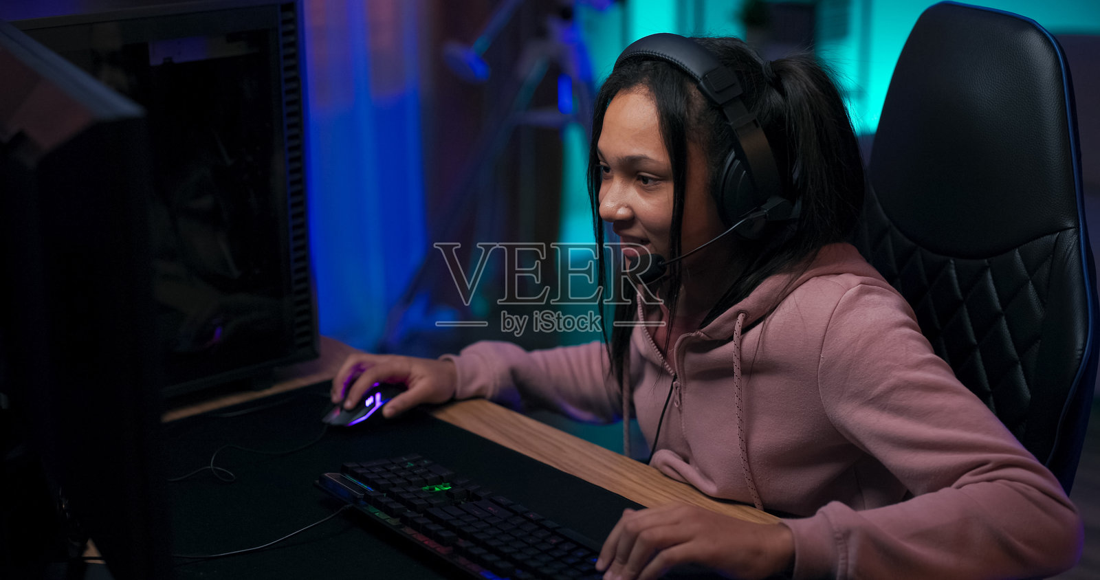 热爱虚拟世界的少女在电脑上玩射击游戏，兴奋地用手指快速点击键盘，佩戴耳机，房间内有蓝色灯光的背光配件。照片摄影图片