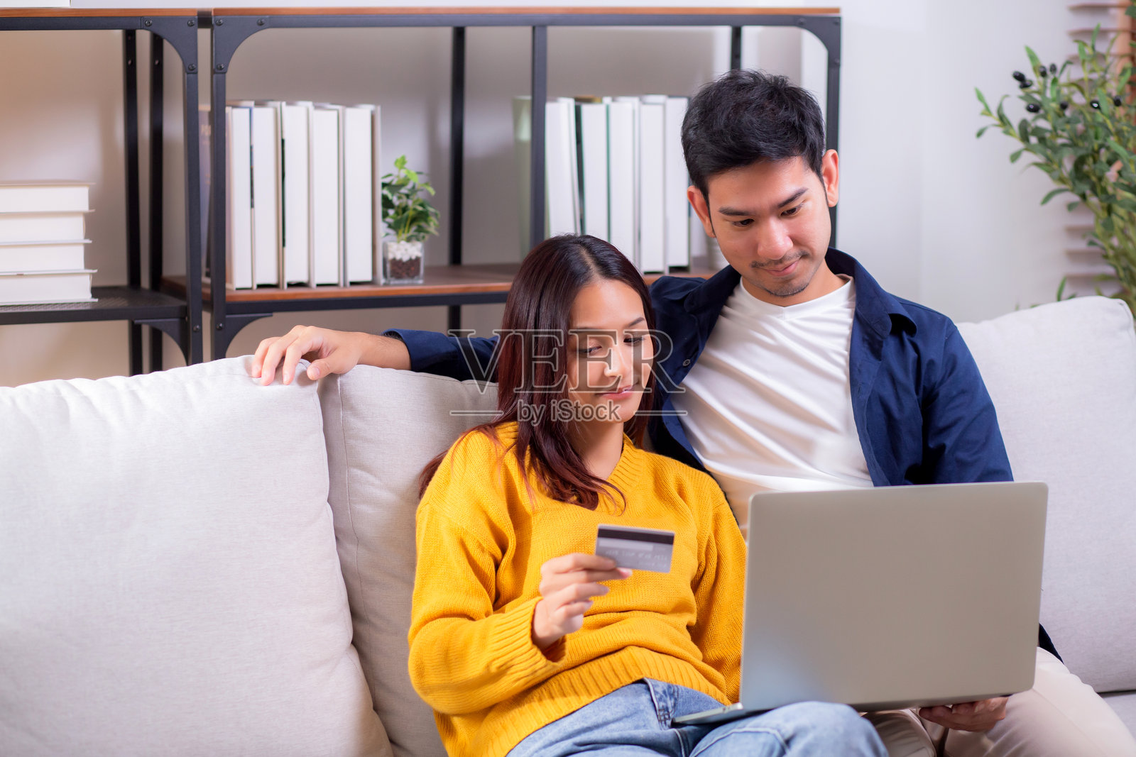 快乐的亚洲情侣在家中的客厅里一起使用笔记本电脑和信用卡进行在线购物，男士和女士微笑放松，享受数字生活方式，便捷的支付体验。照片摄影图片