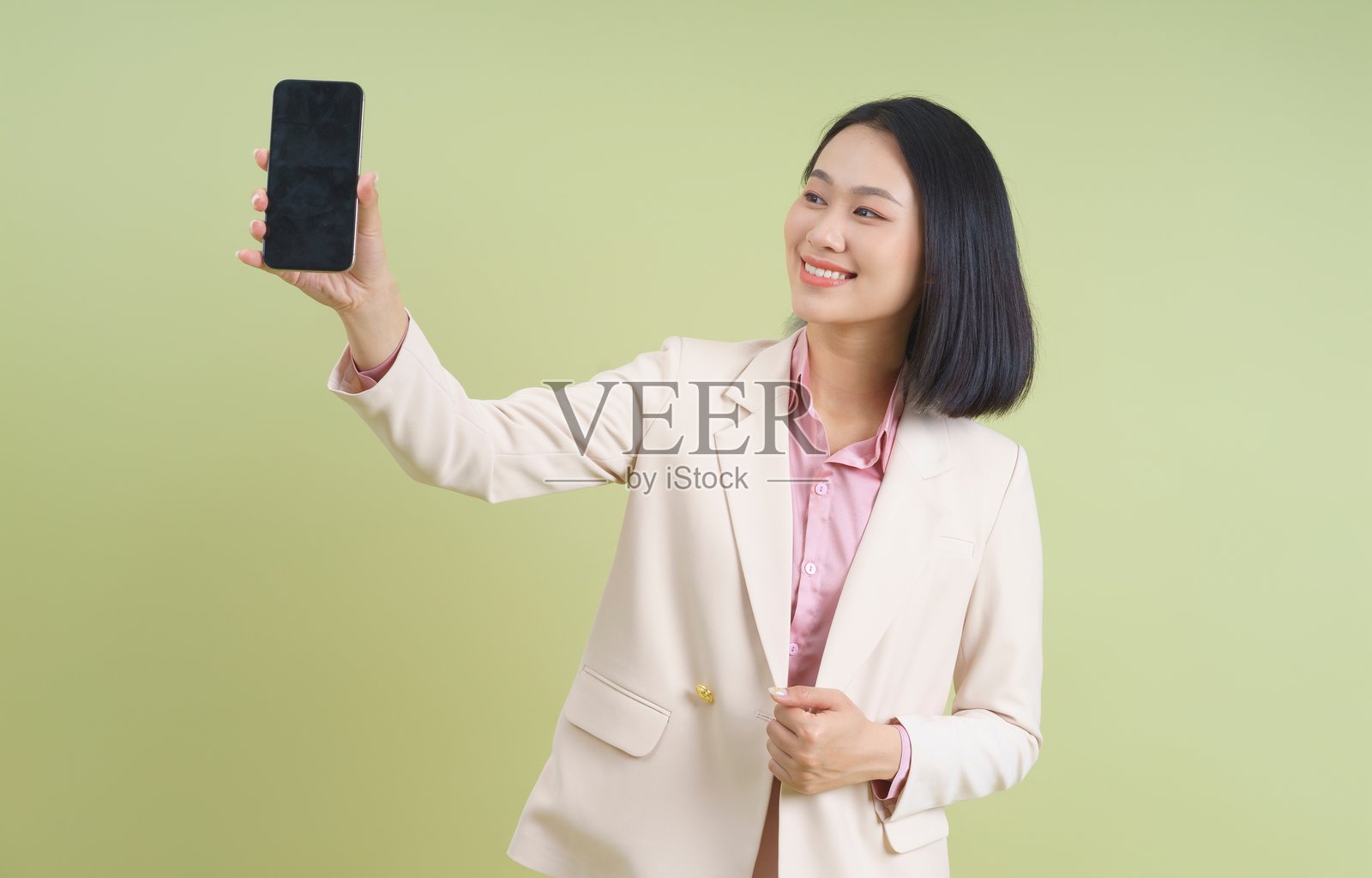 年轻的亚洲女性在休闲商务环境中，微笑着用智能手机在绿色背景前自拍。照片摄影图片